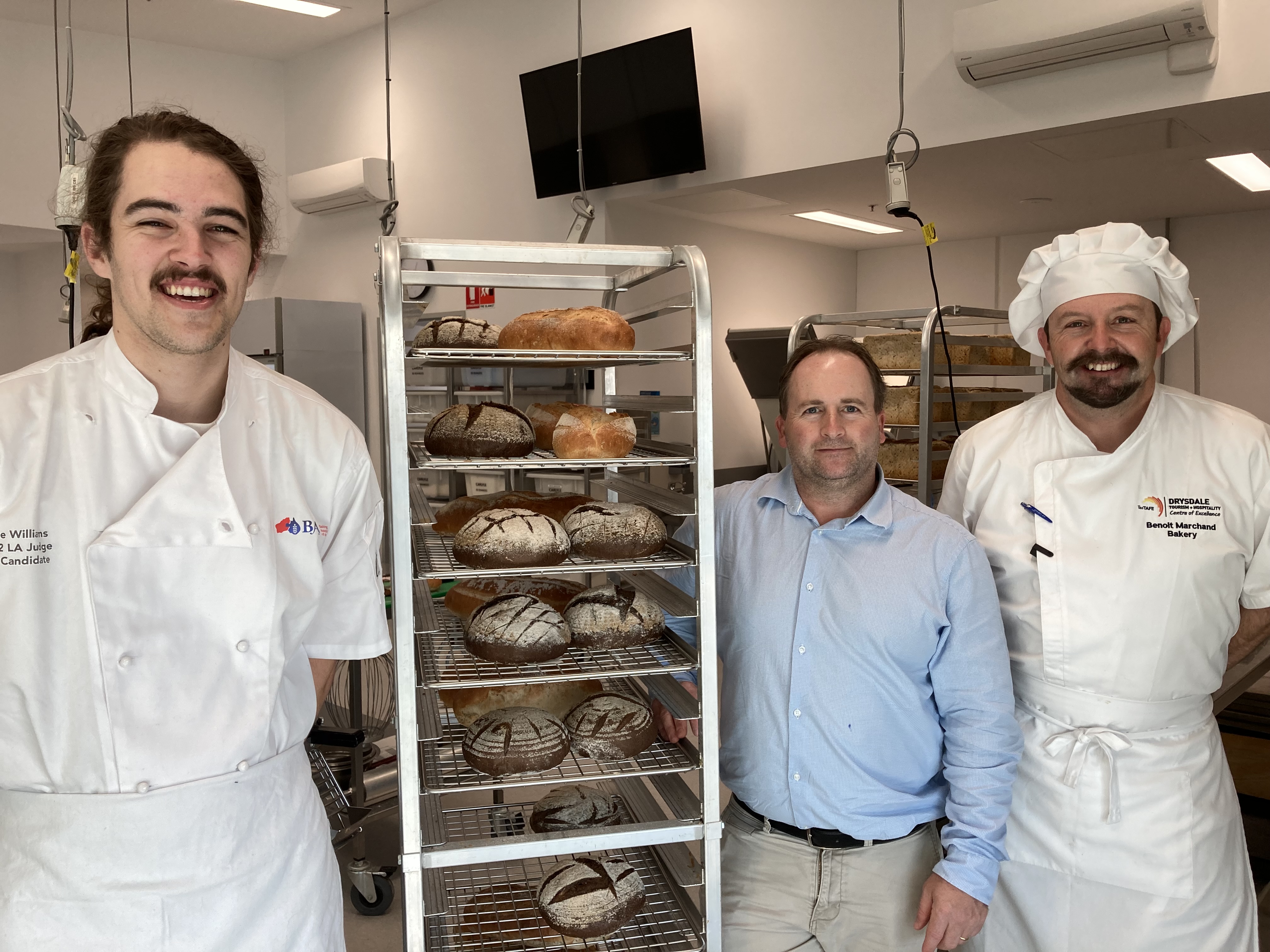TasTAFE apprentice posing with teacher in front of a rack of bread in the baking kitchen