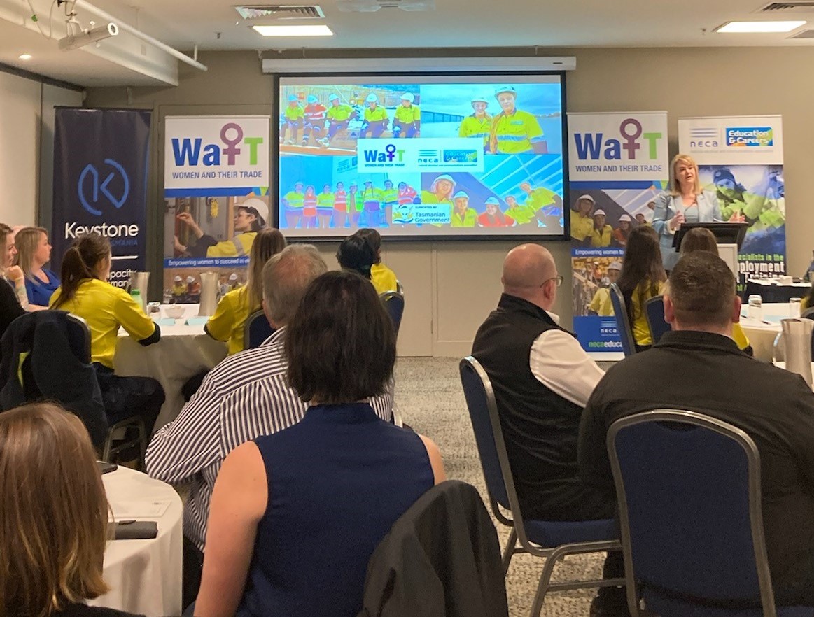 A room full of people sitting around tables at a workshop looking at a presentation on a projector screen