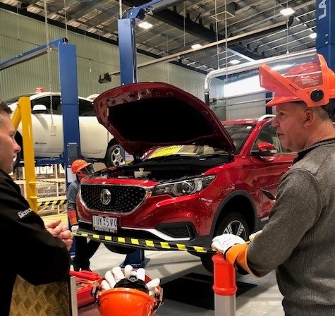 2 people standing in an automotive workshop with a car jacked up with it's bonnet popped in the background