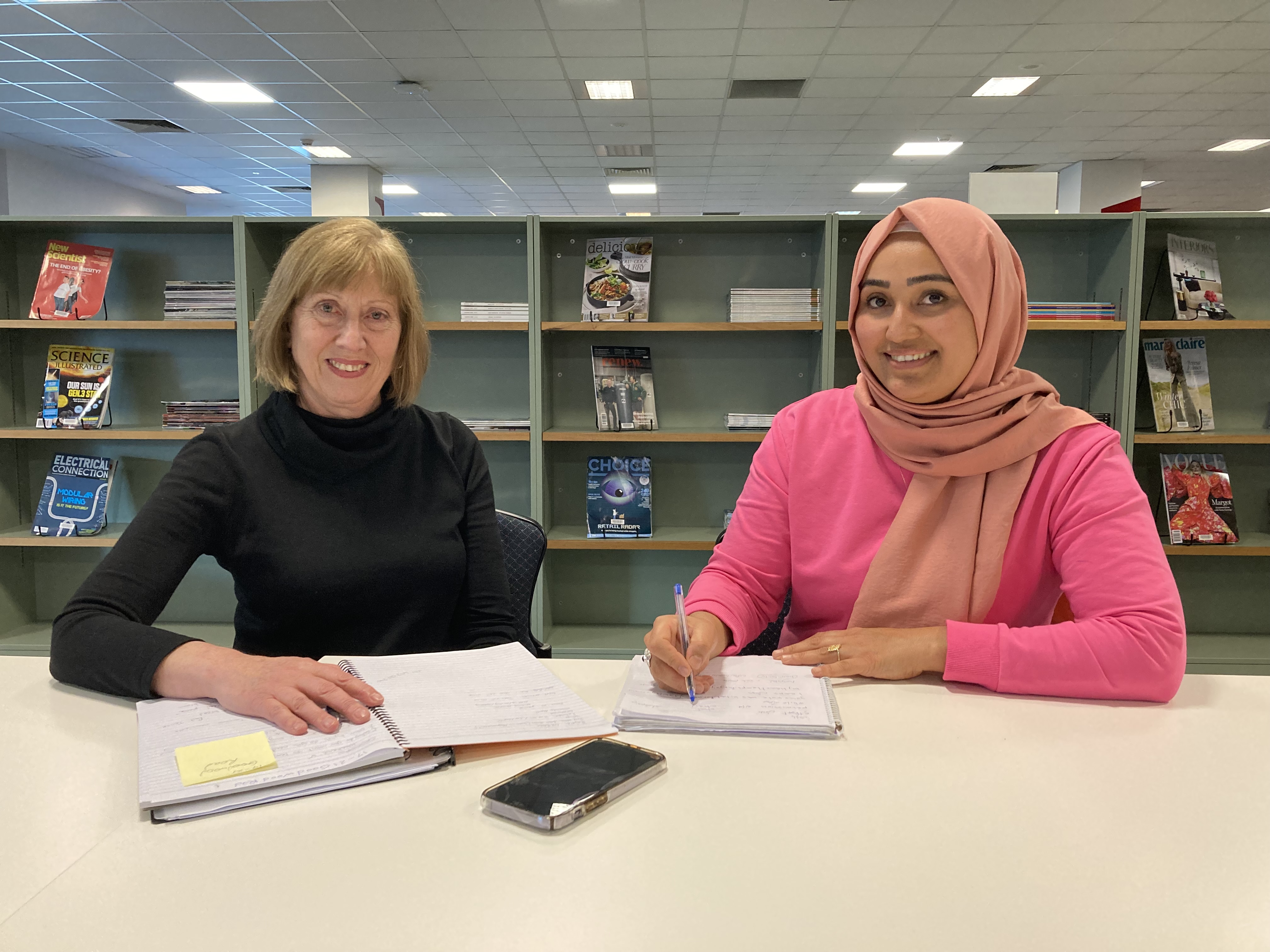 Two women sitting at a desk in the library with notebooks