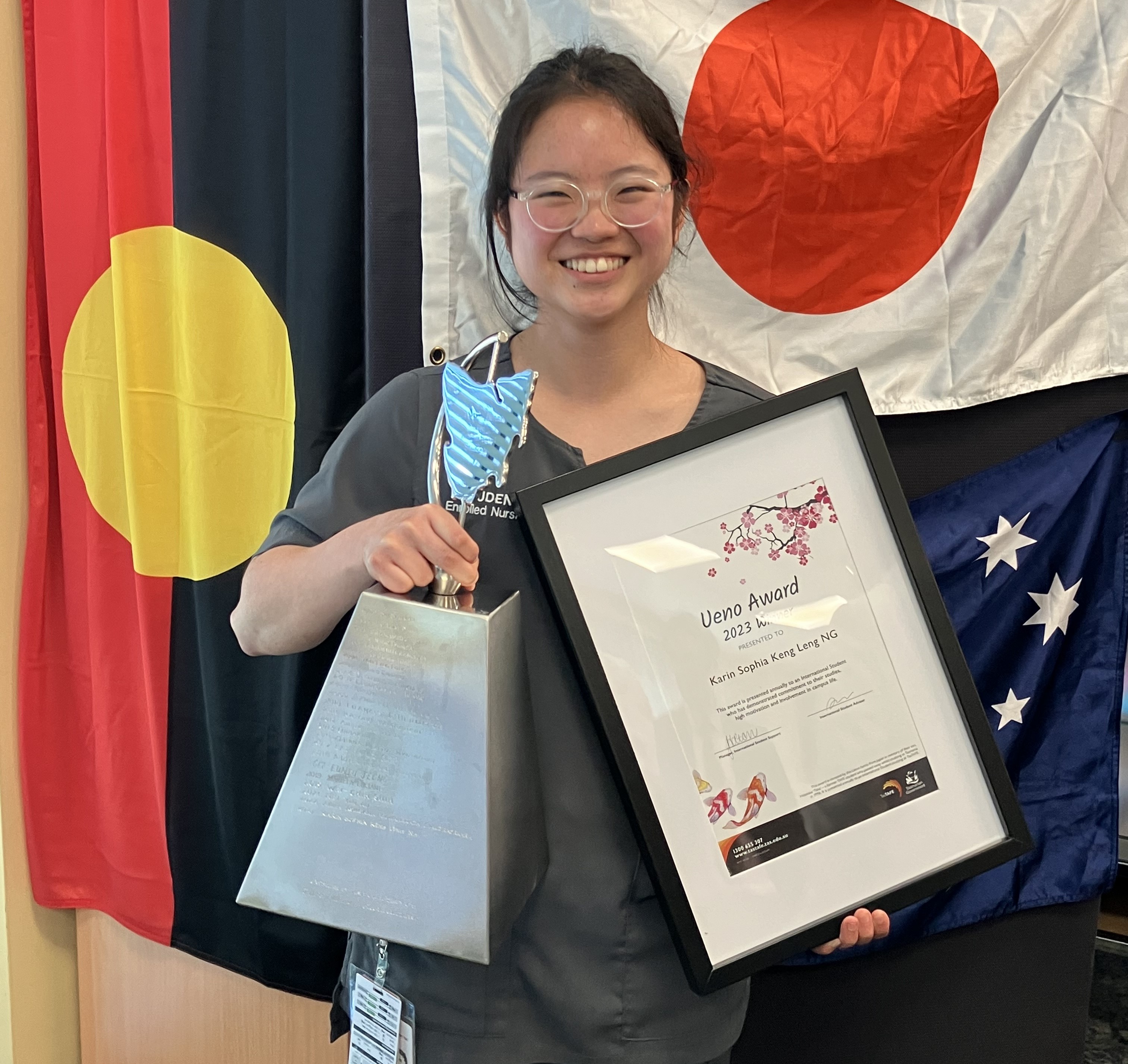 UENO 2023 Award winner Karin Sophi Keng Leng NG holding her certificate in front of the Australian, Japan and aboriginal flags