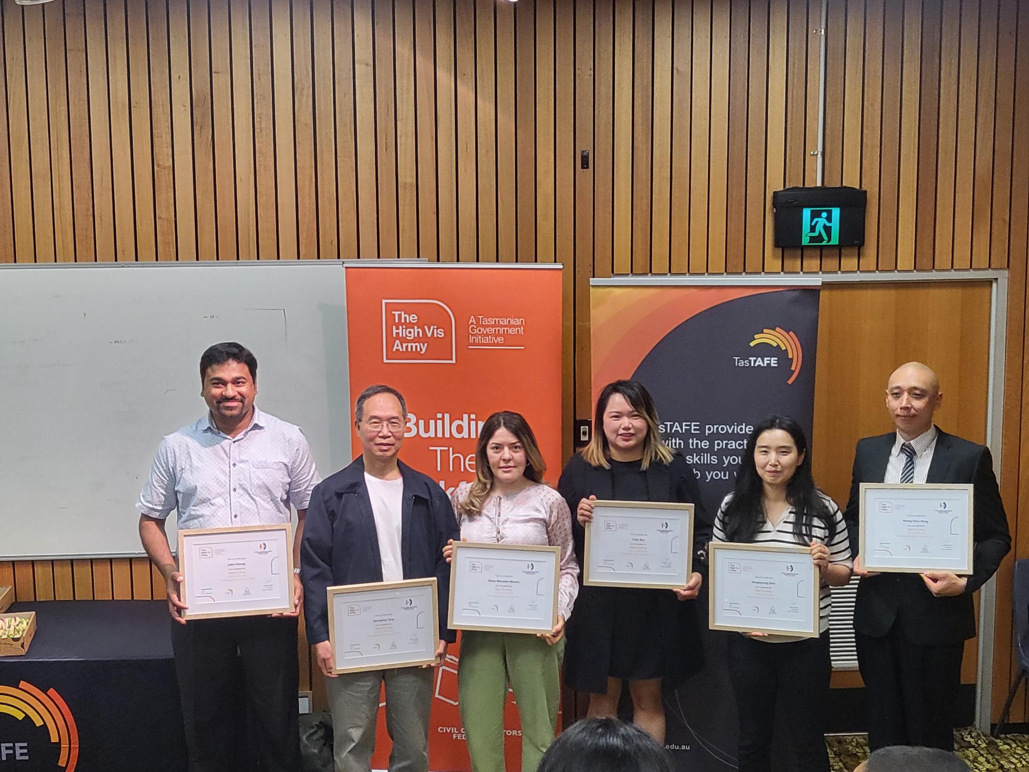 6 people holding their certificates at the front of a lecture theater. 