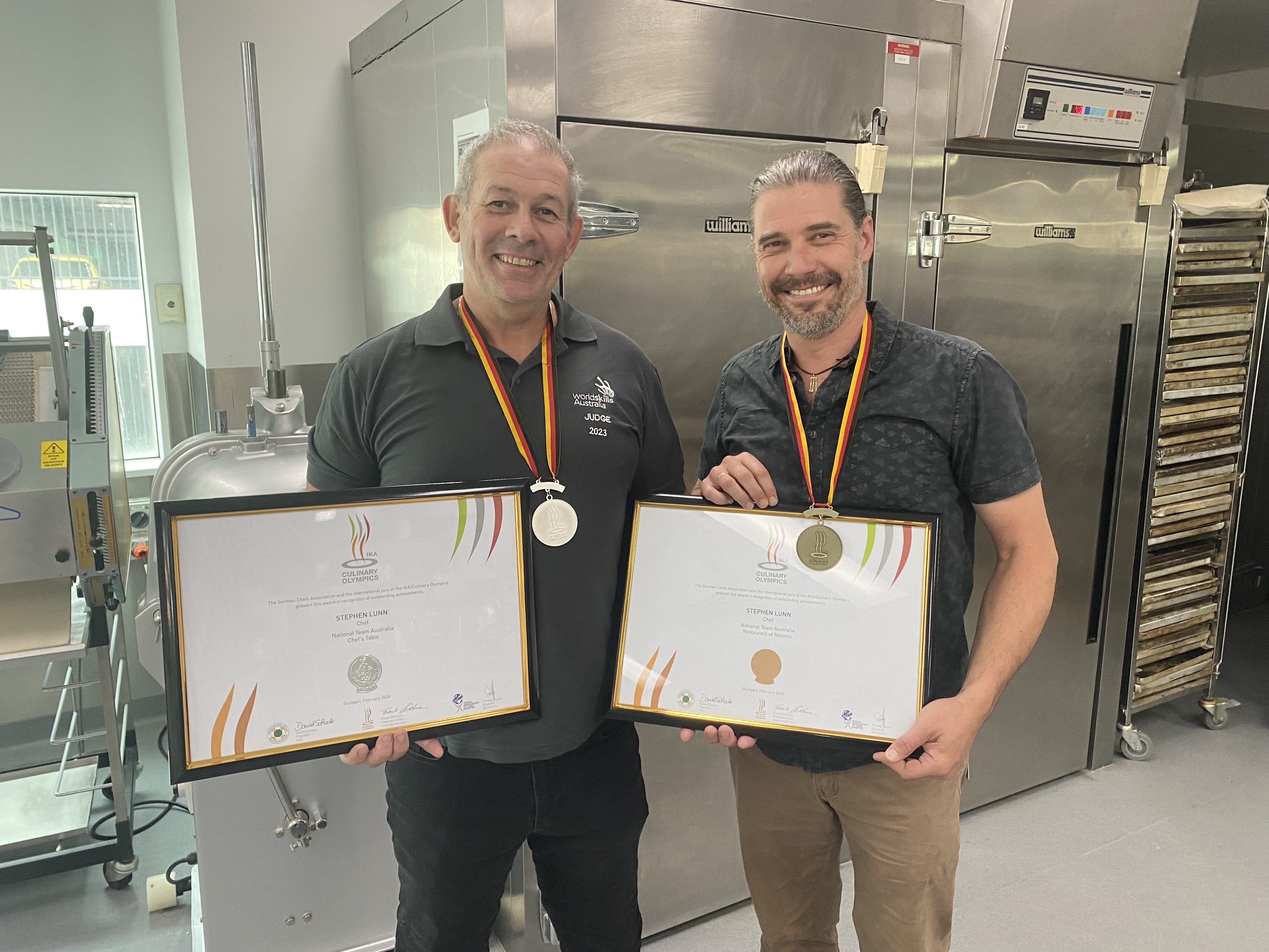 2 men holding framed certificates in the kitchen