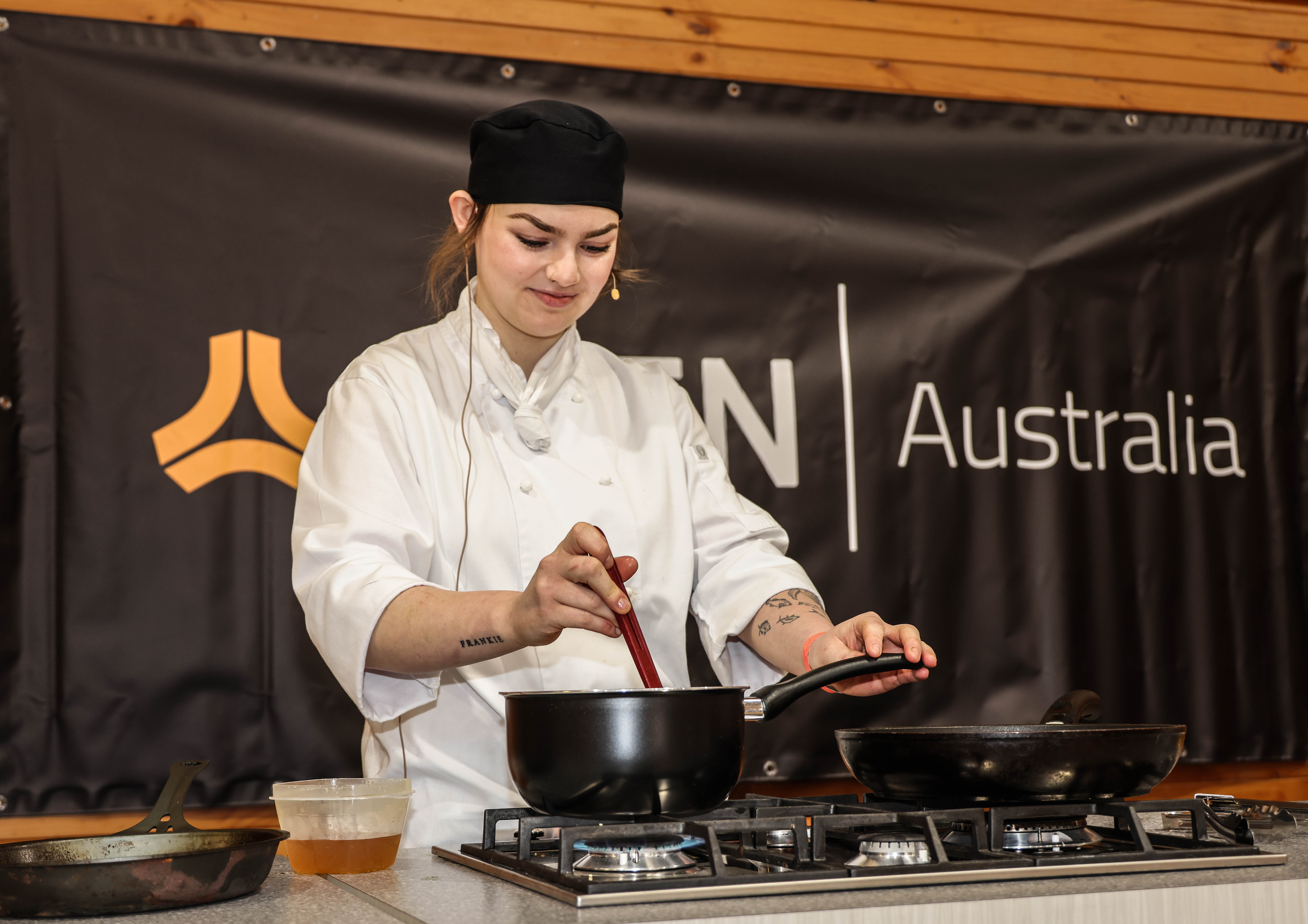 A girl wearing chef's jacket and a black hat, stirring a pan on a gas stovetop conducting a cooking demonstration.