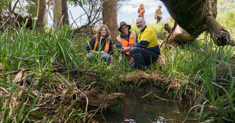3 people crouched in grass in front of water wearing hi vis clothing