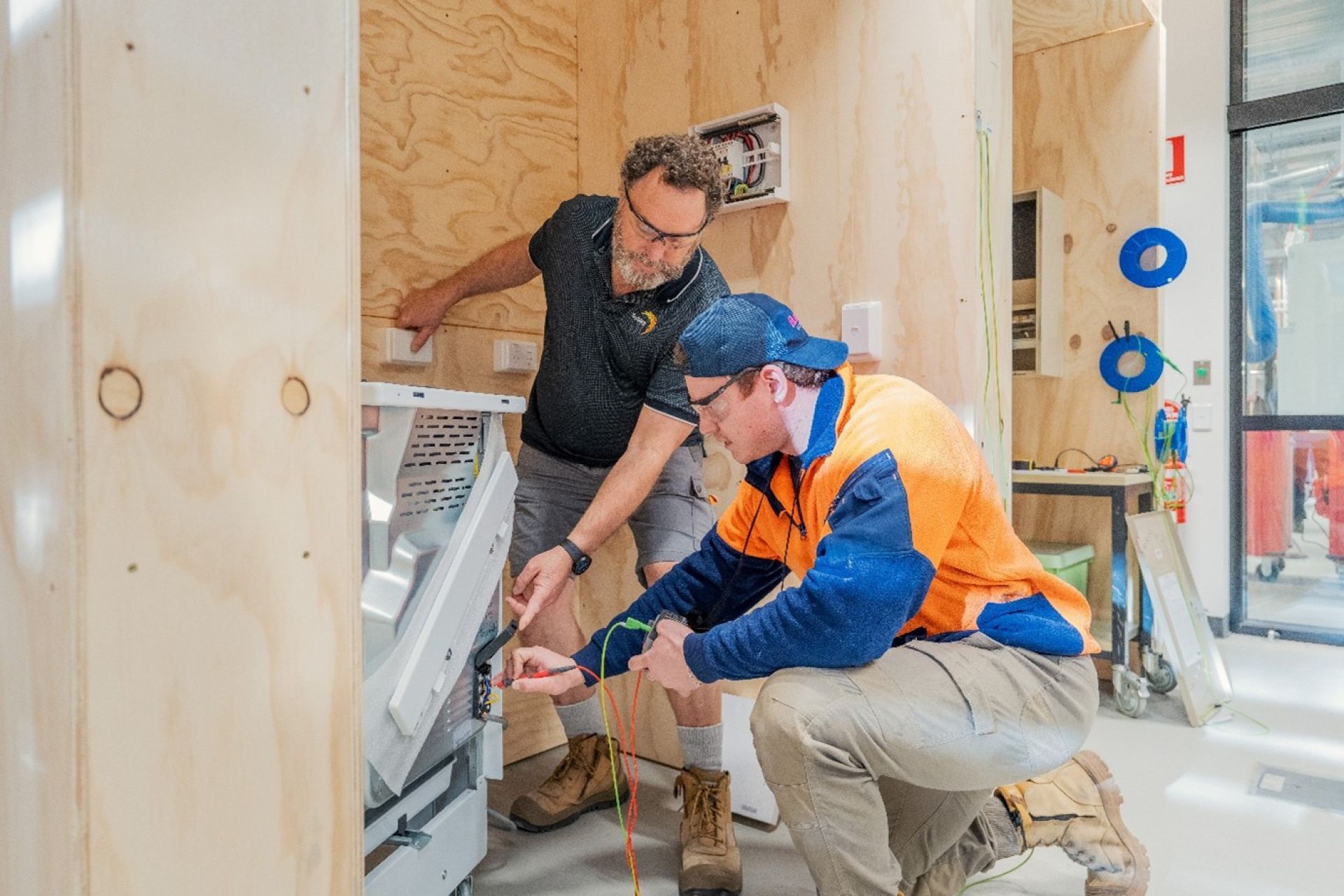 A TasTAFE teacher and a student working together on an electrical installation in a light timber-framed room