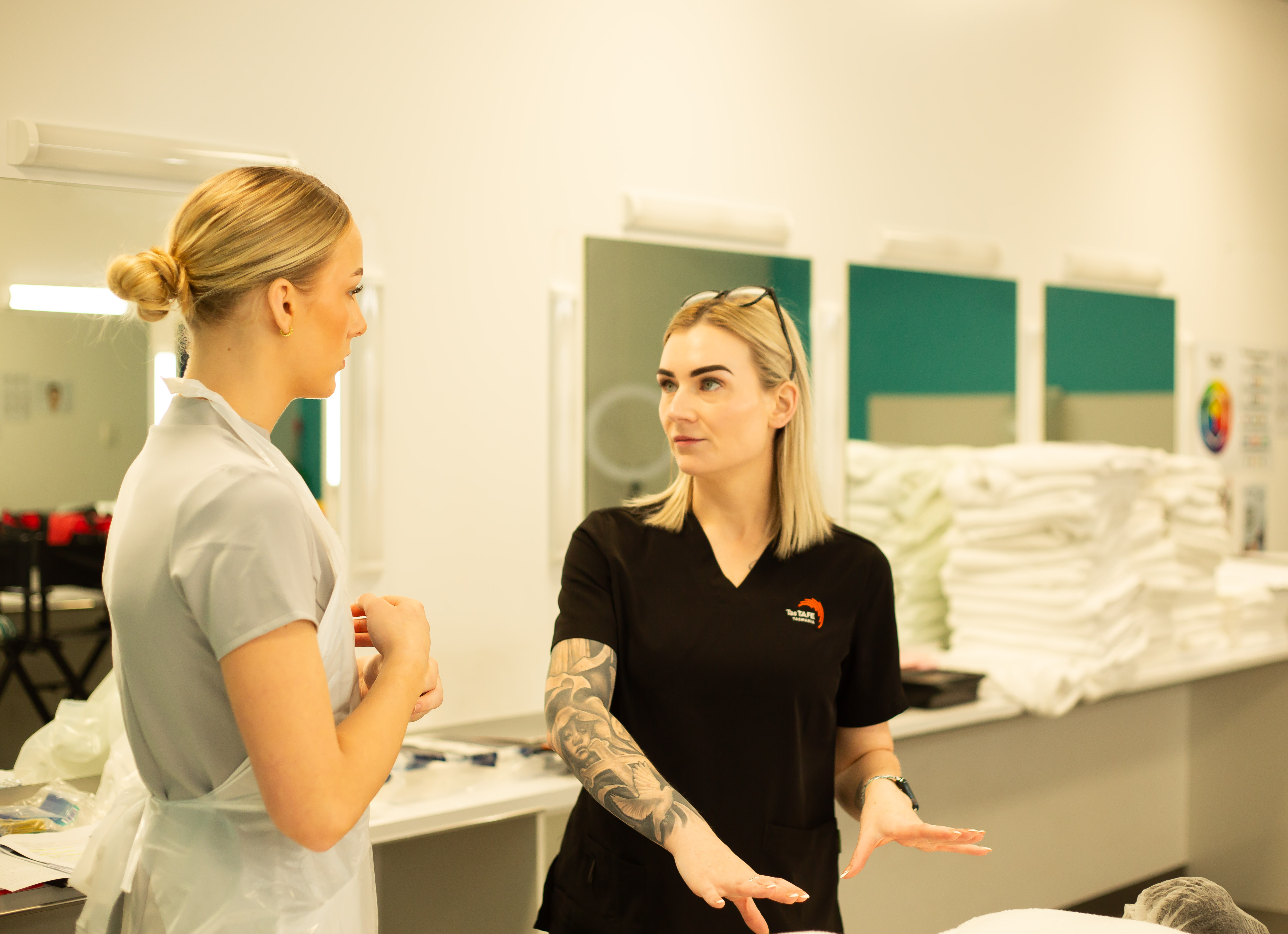 A student and trainer in uniform discussing techniques inside a hair and beauty training salon, with mirrors and stacks of white towels behind them.