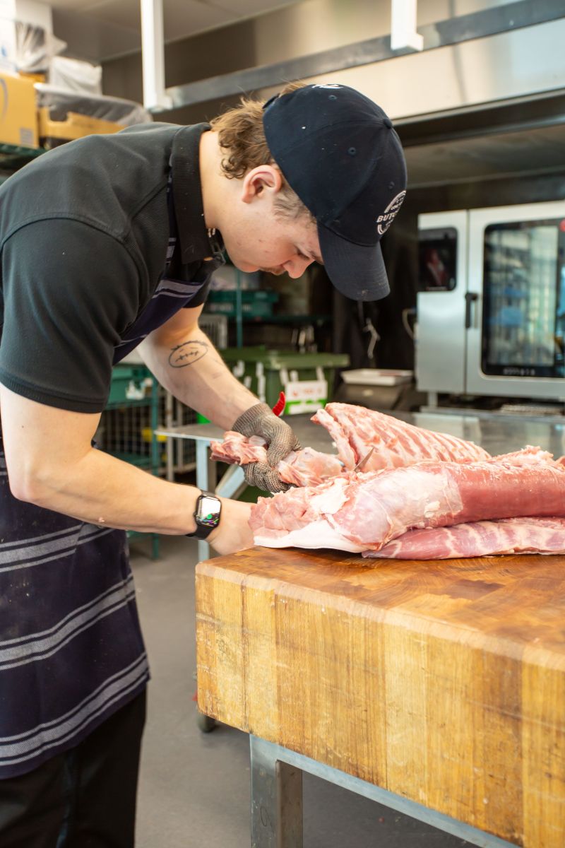 A young butcher wearing a mesh safety glove trims a large cut of raw pork on a thick wooden chopping block in a commercial kitchen.