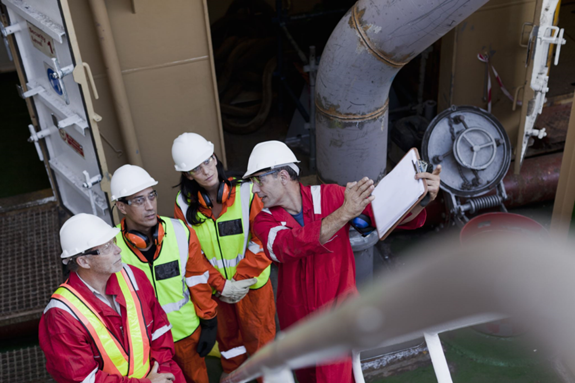 3 people in Hi-Vis clothing listening to an instructor explain something with a clipboard in his hand. 