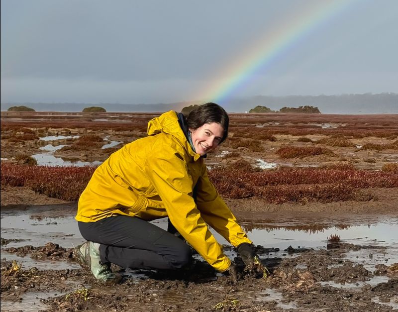 A girl in yellow jacket kneeling and digging in a muddy salt marsh