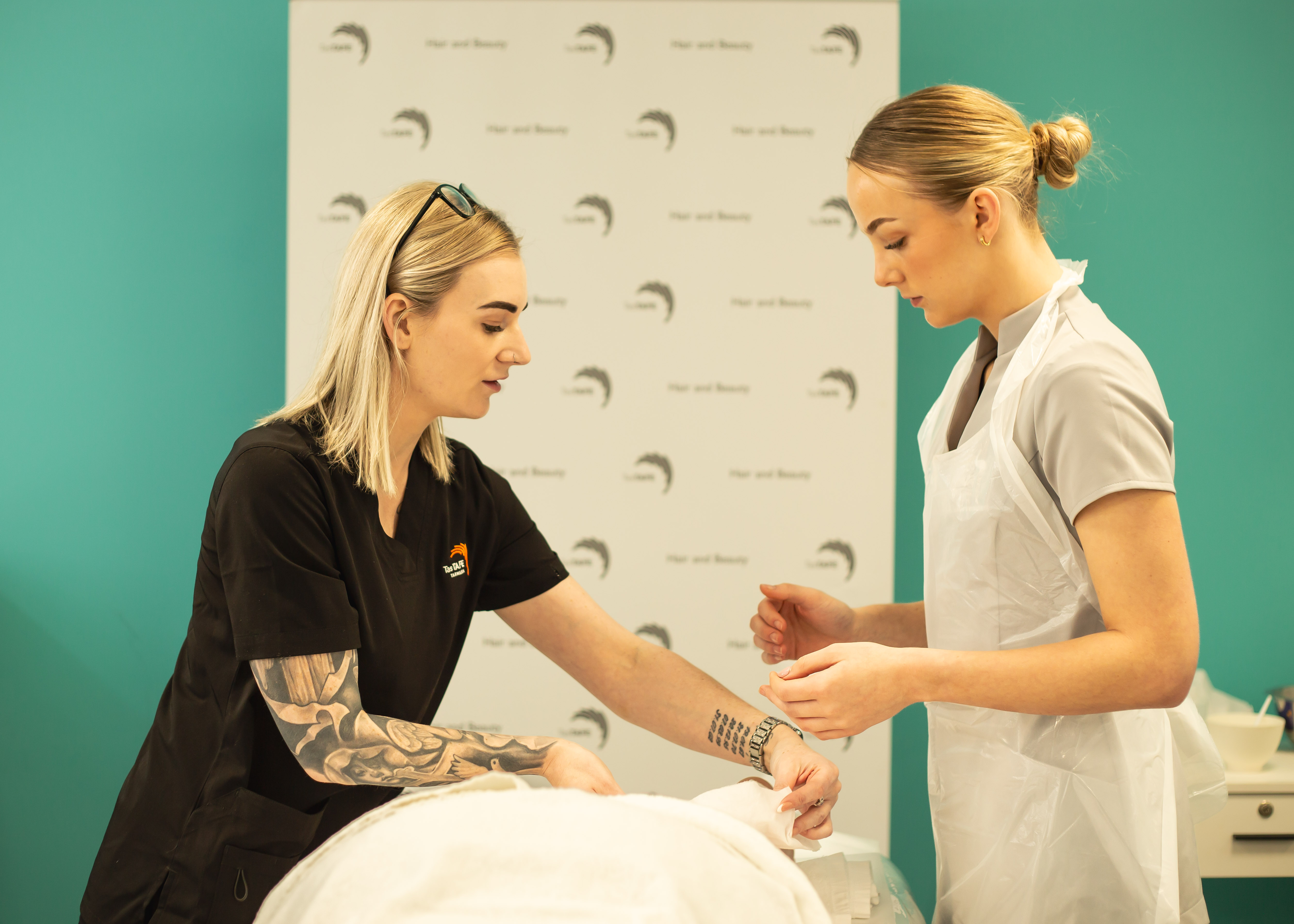 A student in a white apron observes her trainer demonstrating techniques on a treatment table during a practical hair and beauty class.