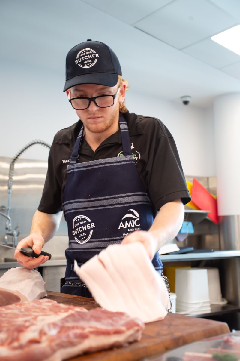 A young butcher with glasses and a cap carefully removing the rind or fat from a piece of raw meat at a wooden workstation.