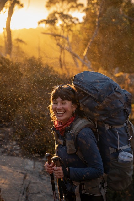 A woman carrying backpacks smiling and walking in the bush
