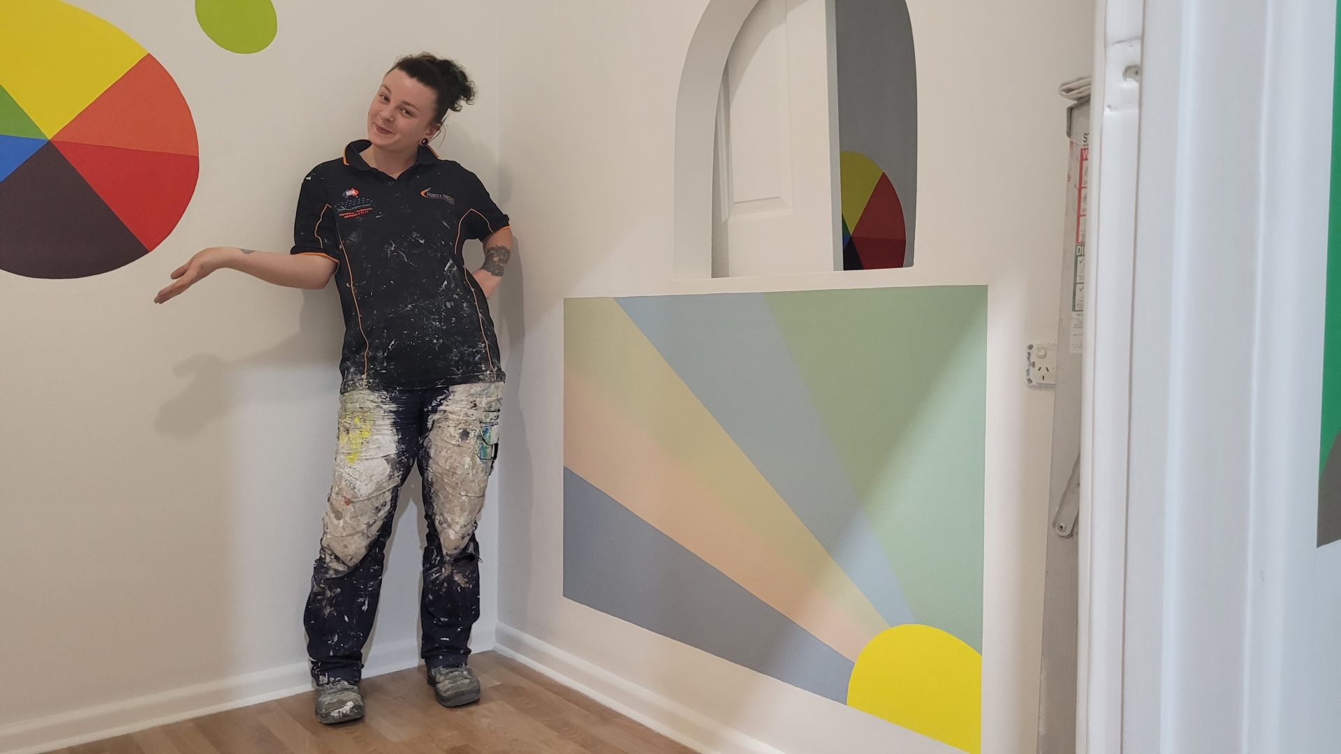 Woman in her paiting clothes in the workshop posing in front of her decorative paintings on the wall