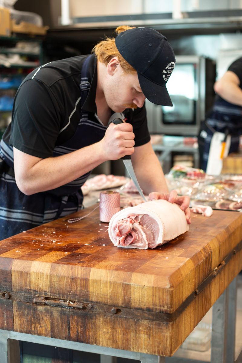 A butcher using a boning knife to prepare a rolled pork roast on a large wooden block, with a spool of butcher's twine nearby.
