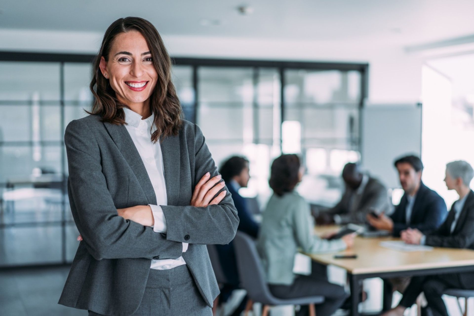 A smiling woman in a suit standing arms crossed with a team meeting in the background