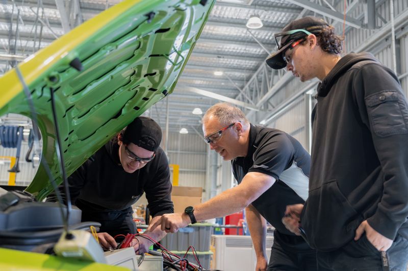 The trainer demonstrating vehicle maintenance techniques to two students in a workshop environment 