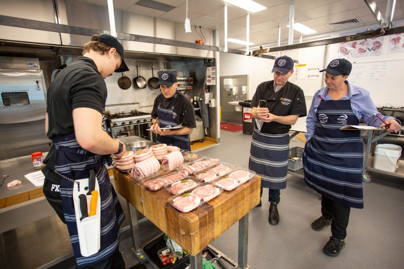 Four butchers in a commercial kitchen wearing striped aprons, with one prepping fresh pork cuts on a wooden block while others judge and take notes.