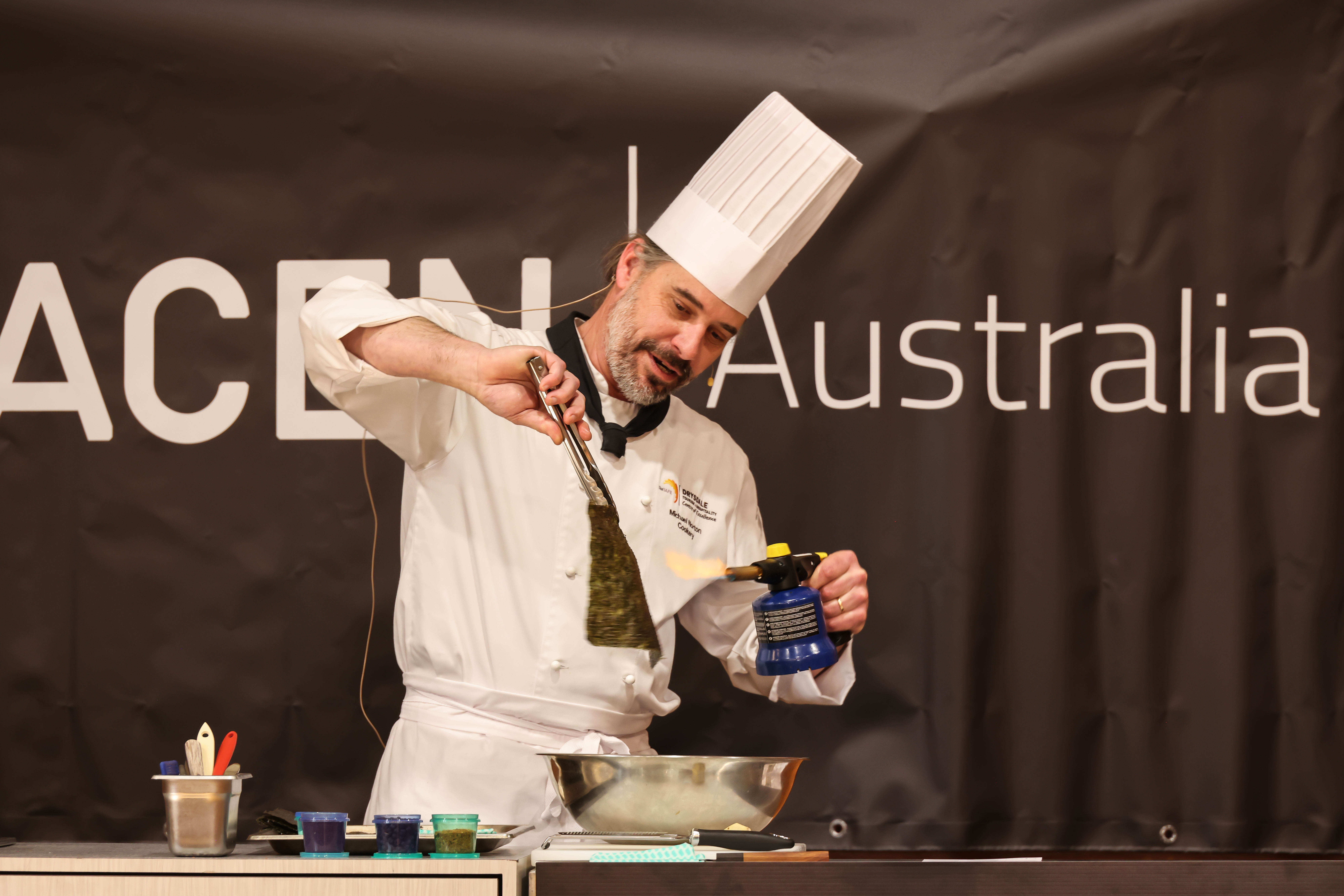 A chef wearing traditional white chef's uniform and demonstrating blowtorch technique on food.
